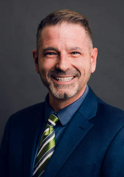Nic Arters in a navy suit in front of a grey background smiling at the camers
