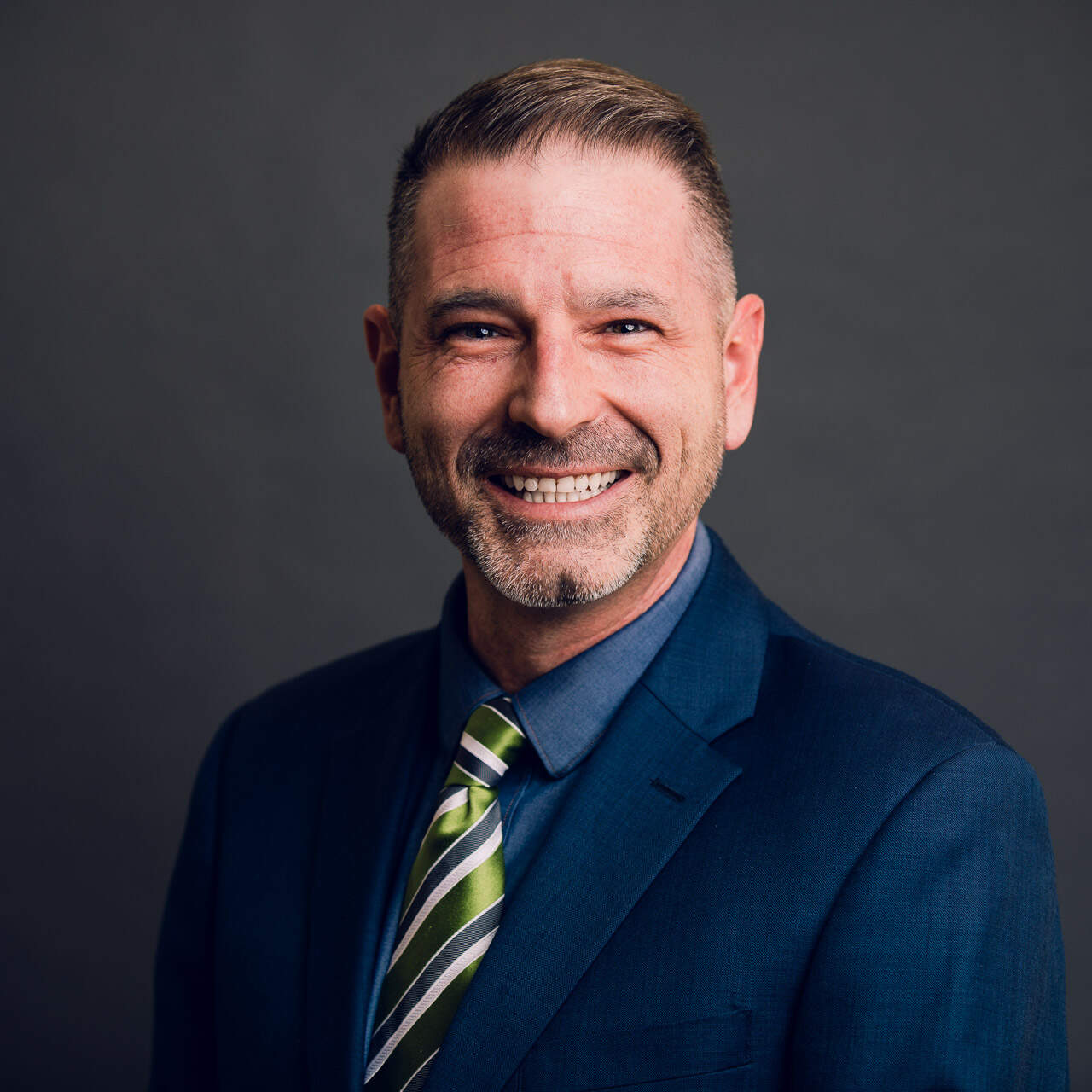 Nic Arters in a navy suit in front of a grey background smiling at the camers